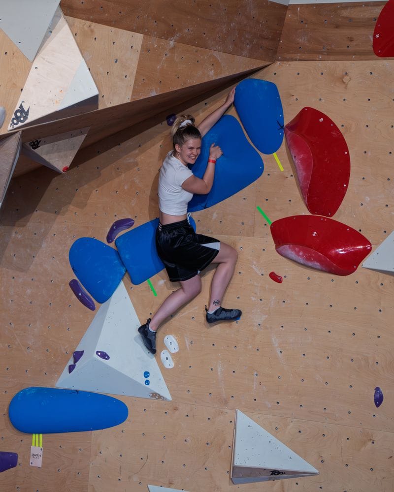Une grimpeuse sourit en plein bloc sur des volumes bleus